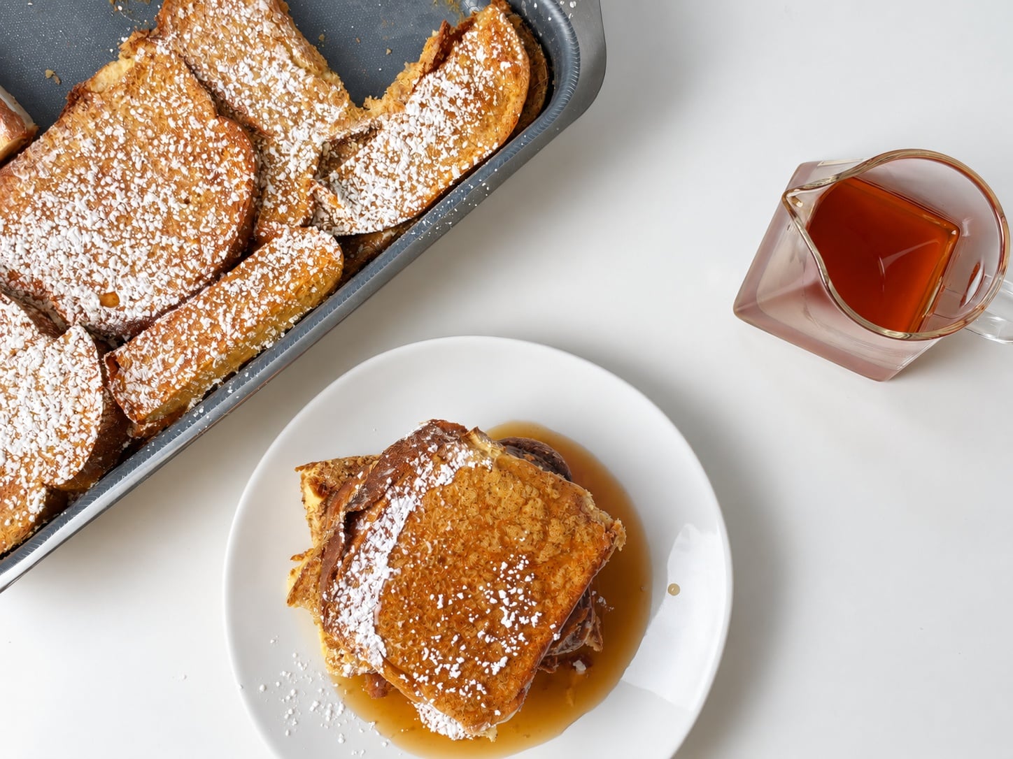 Overhead view of a plated French toast casserole serving with syrup beside the baking pan and syrup pitcher.