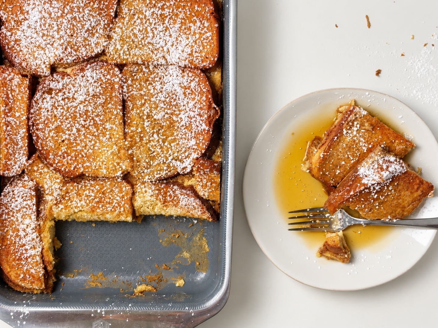 Two pieces of French toast casserole on a plate with syrup beside the partially served baking pan.