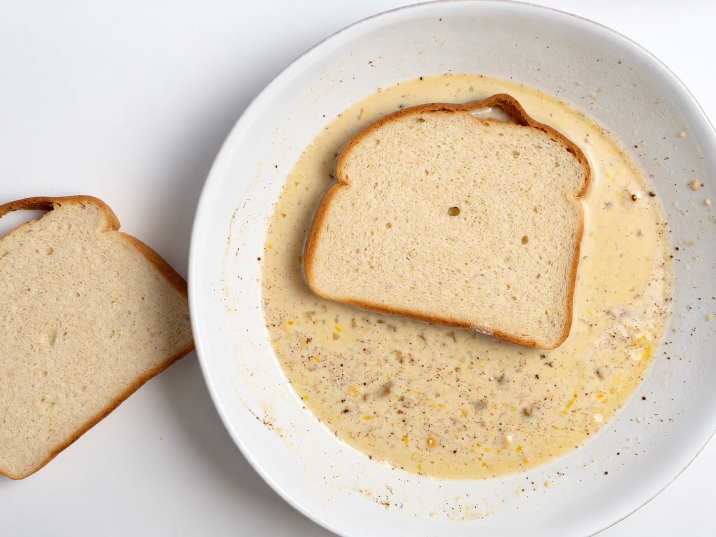Slice of protein bread soaking in cinnamon egg and protein custard in a shallow bowl.