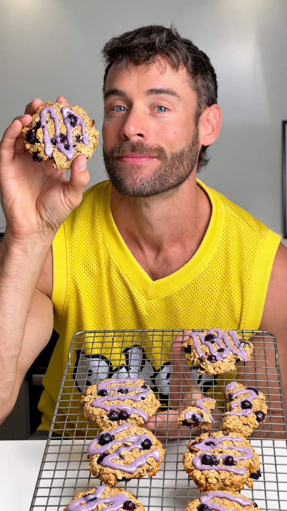 Jacob holding a glazed high-protein blueberry breakfast cookie beside a tray of cookies