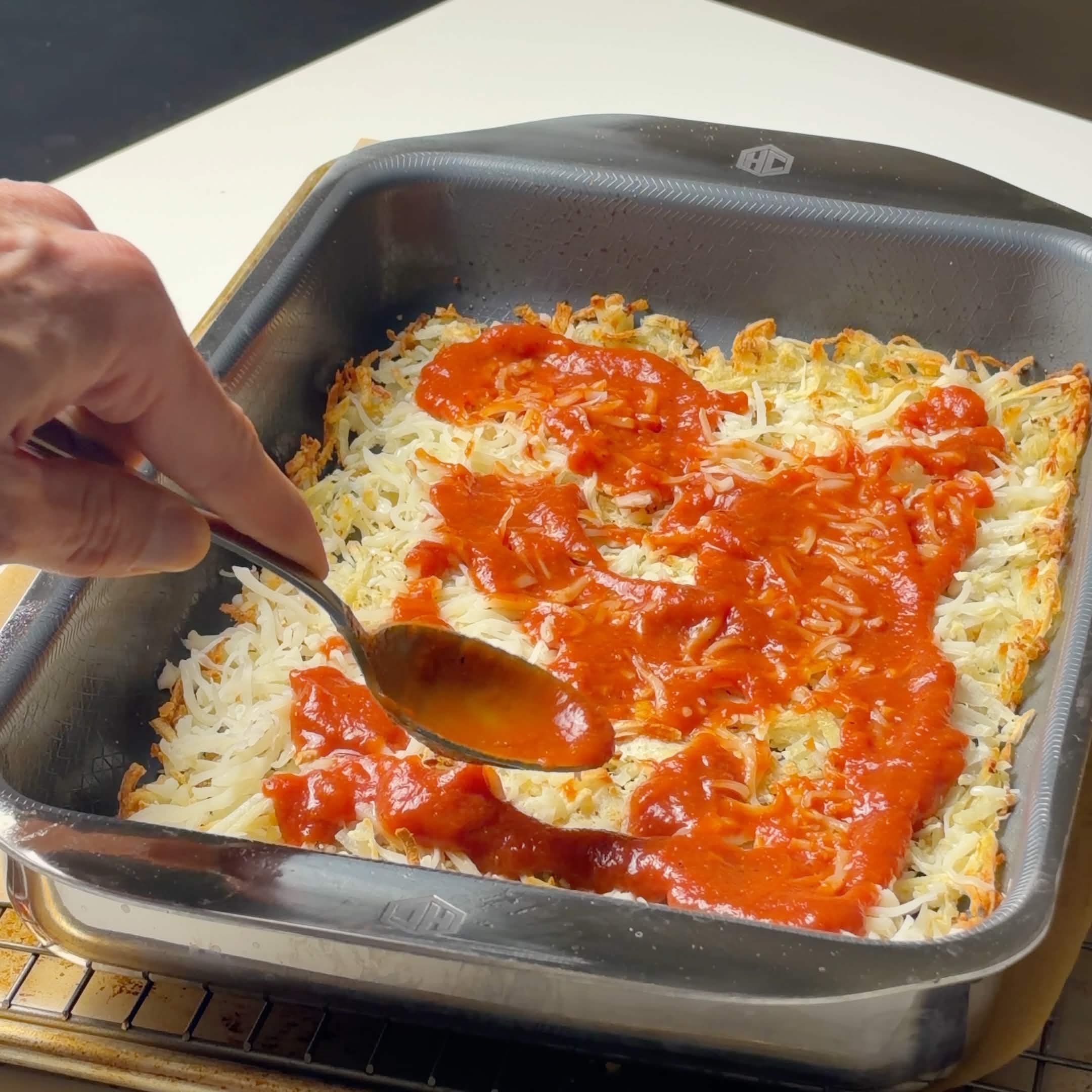 Pizza sauce being spread over a baked shredded hash brown pizza crust