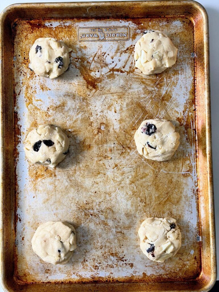 Six large white chocolate cherry almond cookie dough portions spaced on a well-used baking sheet.