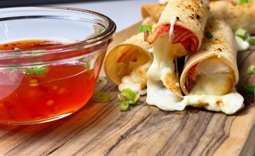 Close-up of crab rangoon mozzarella taquitos filled with imitation crab and melted cheese beside sweet chili sauce.