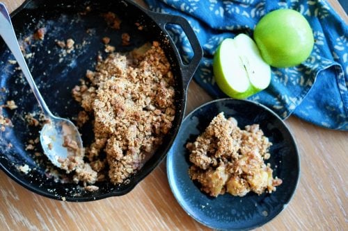 Overhead image of apple crisp in a mini cast iron skillet next to sliced green apples on a wooden table.
