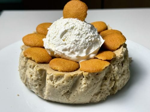 Banana cream cheesecake topped with whipped cream and vanilla wafer cookies on a white plate.